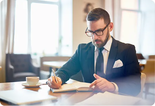 man in a professional setting, likely a cafe or office, writing in a notebook.